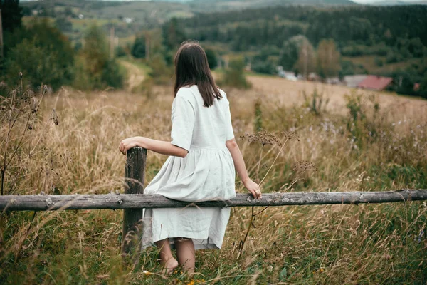 Stijlvolle meisje in linnen jurk zittend op verouderde houten hek onder h — Stockfoto