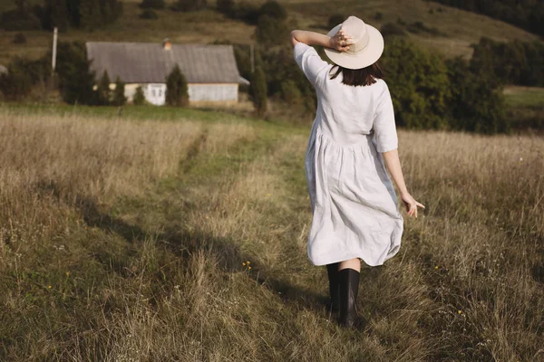 Stylish girl in linen dress and hat walking in sunny field grass — Stock Photo, Image