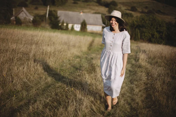 Stylish girl in linen dress and hat walking barefoot in grass in — Stock Photo, Image