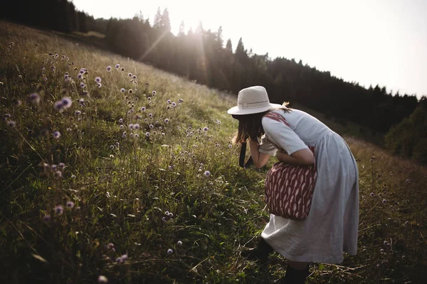 Chica con estilo en vestido rústico y sombrero tomando fotos de flores silvestres —  Fotos de Stock