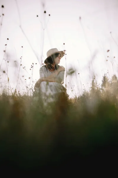 Стильная девушка в ржавом платье и шляпе, сидящая среди полевых цветов — стоковое фото