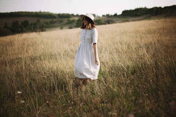 Stylish girl in linen dress walking among herbs and wildflowers in meadow. Boho woman relaxing in countryside, simple slow life style. Atmospheric image. Space text