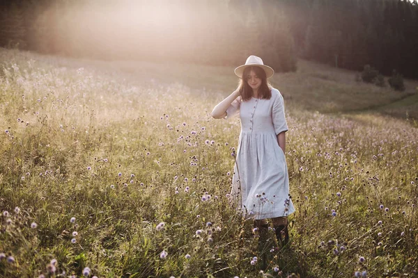 Stylish girl in rustic dress and hat walking among wildflowers i — Stock Photo, Image