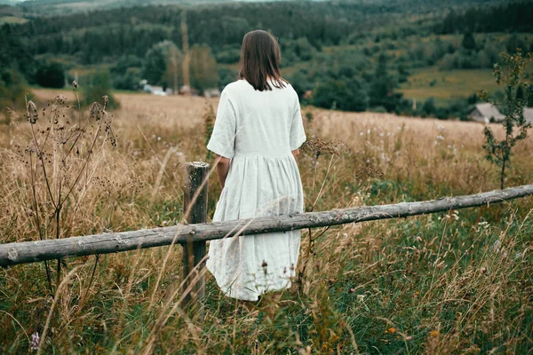 Stylish girl in linen dress sitting on aged wooden fence among h — Stock Photo, Image