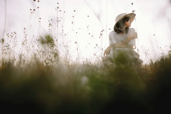 Chica con estilo en vestido rústico y sombrero sentado entre flores silvestres a — Foto de Stock