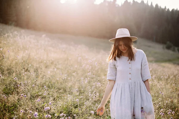 Menina elegante em vestido rústico e chapéu andando entre flores silvestres i — Fotografia de Stock