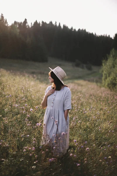 Stylish girl in rustic dress and hat walking among wildflowers a