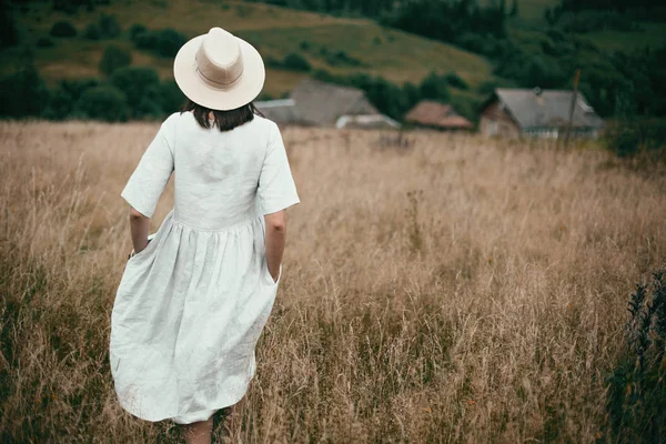 Стильная девушка в льняном платье и шляпе ходит среди трав и диких растений — стоковое фото