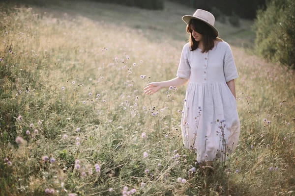 Stylish girl in rustic dress and hat walking among wildflowers a — Stock Photo, Image