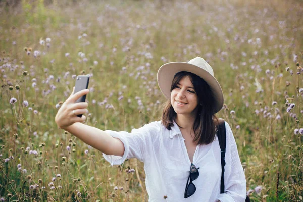 Счастливая девушка-хипстер делает селфи по телефону среди солнечных полевых цветов — стоковое фото