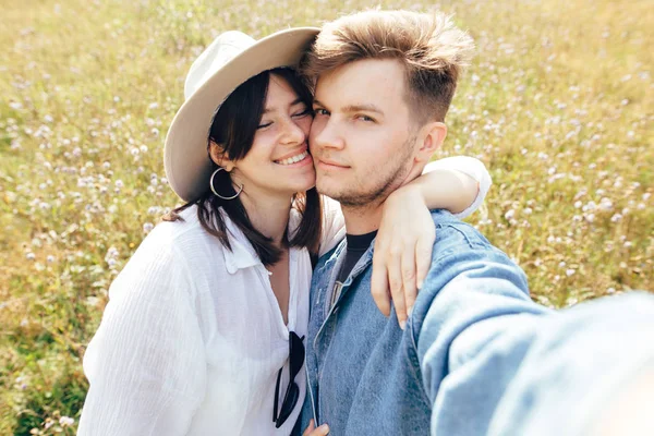 Feliz pareja hipster haciendo selfie en el soleado prado de flores silvestres, t — Foto de Stock