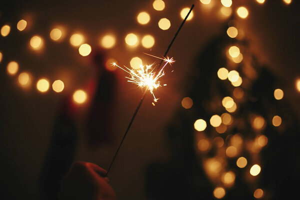 Glowing sparkler in hand on background of golden christmas tree lights, celebration in dark festive room. Happy New Year party. Space for text. Fireworks burning in hand. Happy Holidays