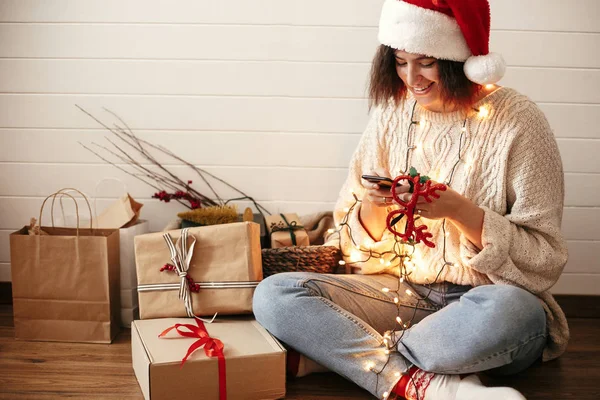 Stylish happy girl in santa hat looking at phone screen in festi — Stock Photo, Image