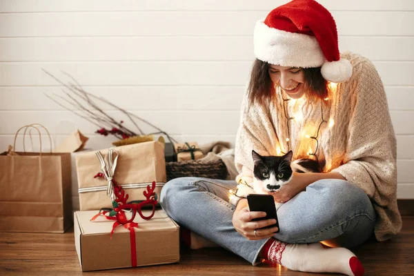 Stílusos boldog lány Santa kalap aranyos macska nézi telefon s — Stock Fotó