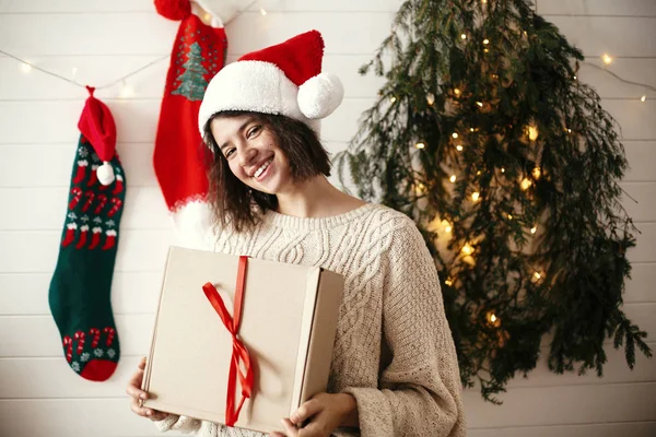 Stylish happy girl in santa hat holding christmas gift box on ba — Stock Photo, Image