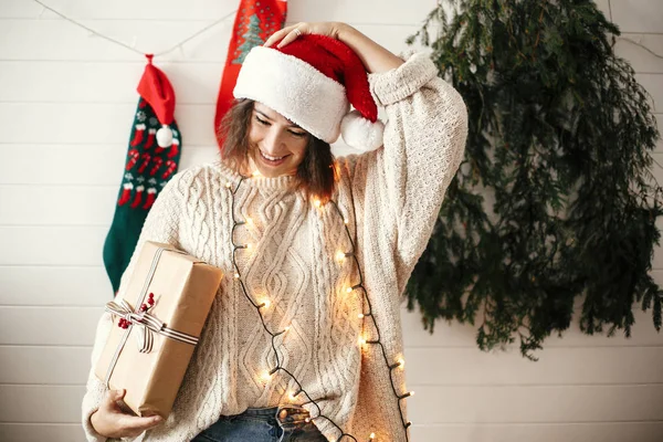 Stylish happy girl in santa hat and cozy sweater holding christm — Stock Photo, Image