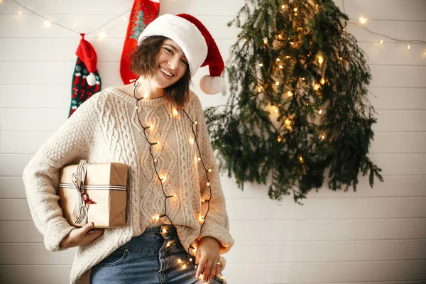 Stylish happy girl in santa hat holding christmas gift box on ba — Stock Photo, Image