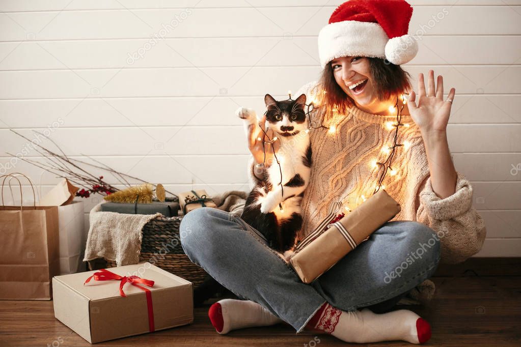 Stylish happy girl in santa hat  and cute cat waving hand and pa