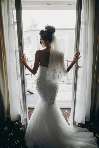 Elegante novia que abre la ventana balcón en luz suave en la habitación del hotel —  Fotos de Stock