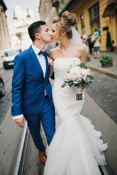 Stylish happy bride and groom walking and kissing in sunny city — Stock Photo, Image