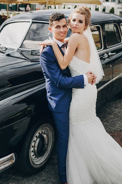 Elegante sposa felice e sposo abbracciando a vecchia auto retrò nero . — Foto Stock