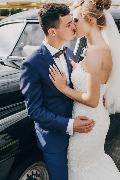 Elegante novia feliz y novio besándose en el viejo coche retro. Precioso. — Foto de Stock