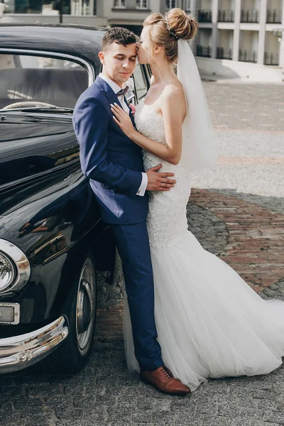 Elegante novia feliz y novio besándose en el viejo coche retro negro. Vamos. —  Fotos de Stock