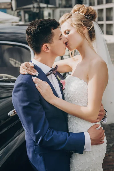 Elegante novia feliz y novio besándose en el viejo coche retro negro. Vamos. — Foto de Stock