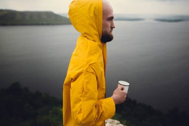 Sarı yağmurluk metal kupa ve trav tutan acımasız sakallı adam