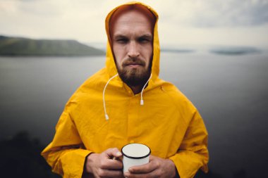 Sarı yağmurluk metal kupa ve trav tutan acımasız sakallı adam