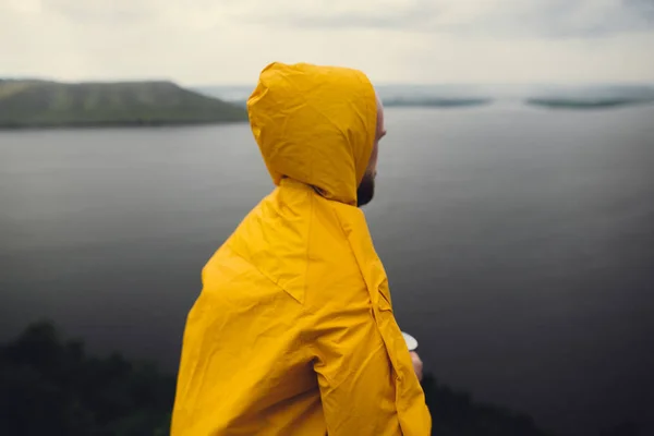 Reisender in gelbem Regenmantel steht auf Klippe und blickt auf See — Stockfoto