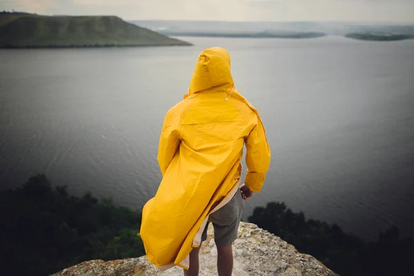 Hipster-Reisender in gelbem Regenmantel steht auf Klippe und blickt — Stockfoto