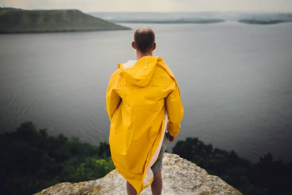 Hipster traveler in yellow raincoat standing on cliff and lookin — Stock Photo, Image