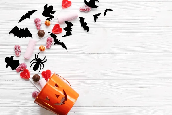 Halloween candy with skulls, black bats, ghost, spider decorations spilled from jack o lantern bucket on white wooden background, flat lay. Halloween sweets. Copy space. Trick or treat