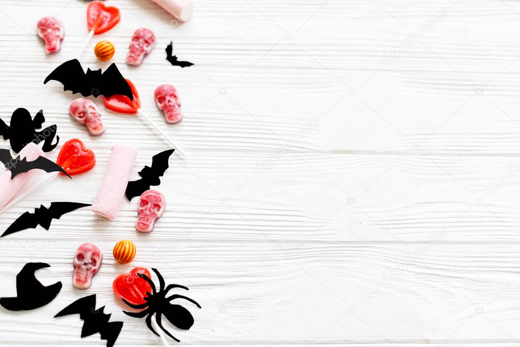 Halloween sweets flat lay. Halloween candy border with skulls, black bats, ghost, spider paper decorations on white wooden background, copy space. Trick or treat concept