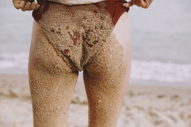 Young woman butt and legs in sand close up on beach. Happy wet girl with sandy swimsuit and legs relaxing on seashore, back view. Summer vacation. Carefree authentic moment.