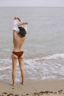 Sahilde ıslak beyaz gömleğini çıkartan mutlu genç bir kadın. Mayolu şık kız deniz kıyısında dinleniyor. Yaz tatili. Kaygısız bir an