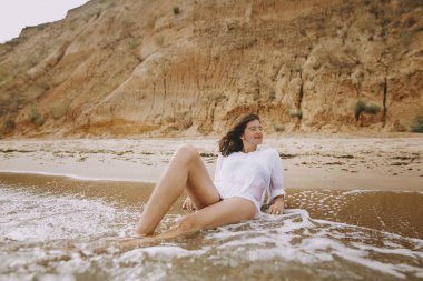 Sahilde dalgalar arasında oturan beyaz tişörtlü mutlu genç kadın. Havalı bronzlaşmış kız deniz kıyısında dinleniyor ve dalgaların tadını çıkarıyor. Yaz tatili. Farkındalık ve kaygısız an