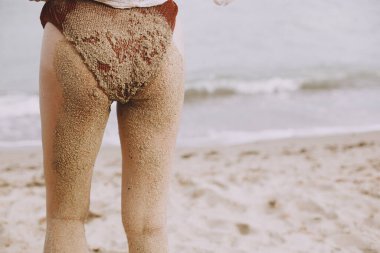 Young woman butt and legs in sand close up on beach. Happy wet girl with sandy swimsuit and legs relaxing on seashore, back view. Summer vacation. Carefree authentic moment.