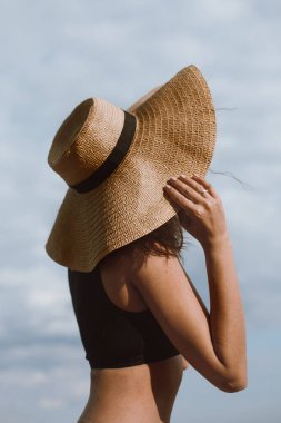 Bronz tenli kız sıcak yaz gününde elinde şapka ve mavi gökyüzünün arka planında poz veriyor. Hasır şapkalı, plajda dinlenen şık bir genç kadın. Kaygısız. Yaz tatili