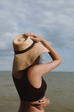 Bronz tenli kız sıcak yaz gününde elinde şapka ve mavi gökyüzünün arka planında poz veriyor. Hasır şapkalı, plajda dinlenen şık bir genç kadın. Kaygısız. Yaz tatili