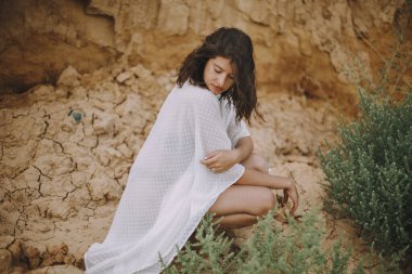 Beyaz yaz elbiseli seksi kız yeşil çalıların arasında kumsalda oturuyor. Güzel estetik bir görüntü. Deniz kıyısında dinlenen genç bir kadın. Yaz tatili. Huzur