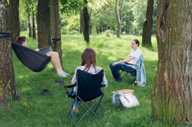 Small group of people enjoying conversation at picnic in accordance with social distancing in summer park. Friends chilling in hammock and on chairs among trees