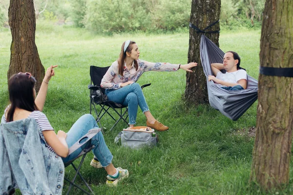 Small group of people enjoying conversation at picnic with social distance in summer park. Social distancing. Friends chilling in hammock and chairs among trees. Lifestyle leisure activity