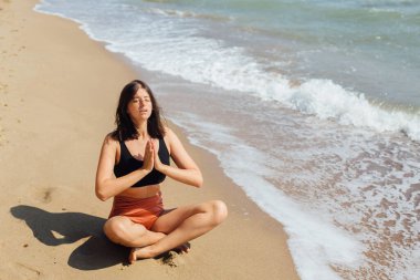Genç ve güzel bir kadın kumsalda yoga yapıyor, kumların üzerinde oturuyor ve meditasyon yapıyor. Akıl sağlığı ve öz bakım kavramı. Mutlu kız yaz tatilinde deniz kıyısında dinleniyor.