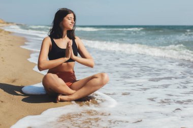Genç ve güzel bir kadın kumsalda yoga yapıyor, kumların üzerinde oturuyor ve meditasyon yapıyor. Akıl sağlığı ve öz bakım kavramı. Mutlu kız yaz tatilinde deniz kıyısında dinleniyor.