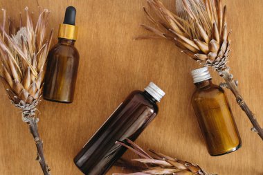 Sıfır atık kozmetik. Şampuan, serum ve ubtan cam şişeler içinde ahşap arka planda kurutulmuş protea çiçekleri, düz yatak. Plastiksiz hijyen ürünleri.