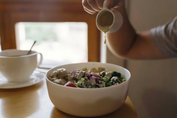 Buddha bowl, healthy eating concept. Man pouring sauce on vegan meal in restaurant. Bowl with avocado, radish, broccoli, tofu, tomato, couscous, microgreen and sesame seeds.