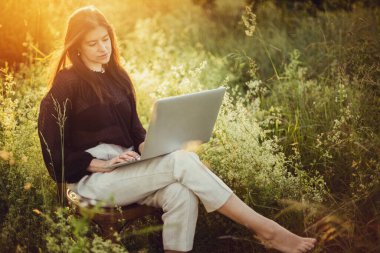 Şık bir kız dizüstü bilgisayarda çalışıyor ve yaz otlağında sıcak gün batımında kırsal sandalyede oturuyor. Sosyal uzaklık ve güvenlik protokolleriyle ilgili yeni çalışmalar. Uzak çalışma