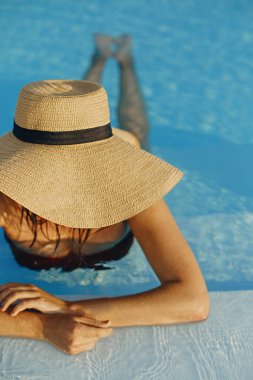 Şapkalı şık genç bir bayan mavi havuzda dinleniyor ve yaz tatilinin tadını çıkarıyor. Yaz tropik tatili. Lüks tatil köyünde güneşli şapkalı bir kız çatıda havuzda yüzüyor. Metin için boşluk